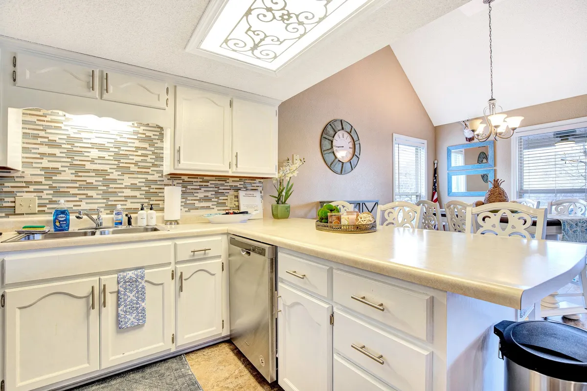 A bright, open kitchen with a central island flows effortlessly into a spacious dining area. Stylish backsplash tiles, a decorative clock, and green plants add warmth and homeliness. The layout supports easy movement and casual gatherings, ideal for sharing meals, games, and stories.