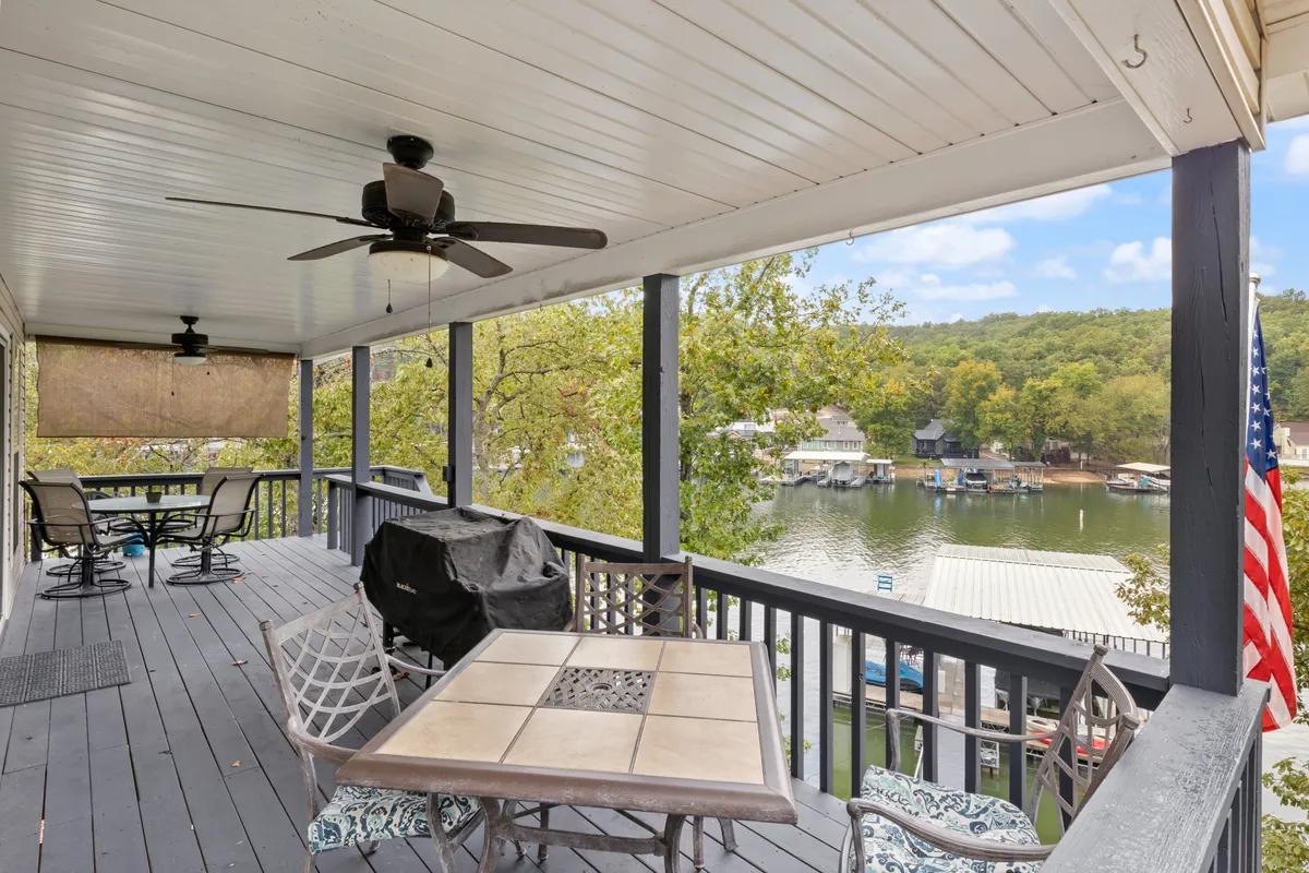 A relaxed lakeside deck scene with an outdoor dining table under a ceiling fan, offering a sweeping view of the lake beyond. The American flag adds a touch of character. The space is set up for both dining and socializing, with ample room and a welcoming atmosphere that fits family meals or evening toasts.