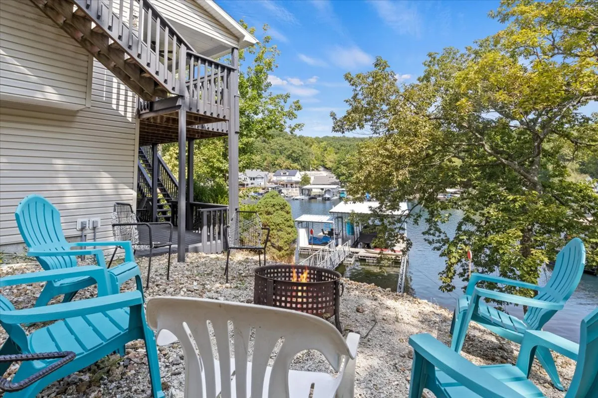 A welcoming lakeside fire pit area with comfortable outdoor seating arranged to enjoy panoramic sunset views over the water. The scene evokes a cozy, social mood, with the fire pit inviting for evening gatherings and s'mores-making, framed by the calm lake backdrop.