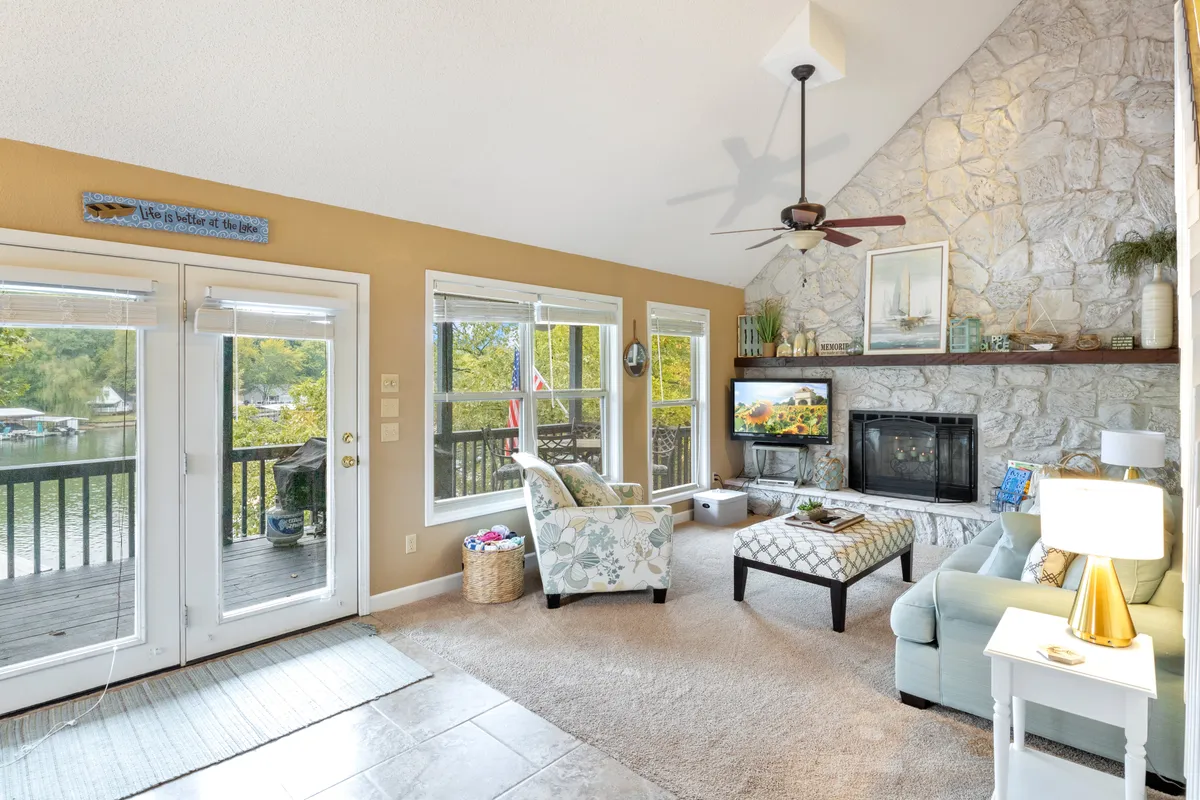 This image features a large stone fireplace as the focal point of a comfortable living area. Plush sofas are arranged to invite group gatherings, with a Smart TV mounted above the mantle. Generous windows showcase serene lake views, and the space is bathed in natural light, enhancing the welcoming, open-concept atmosphere.