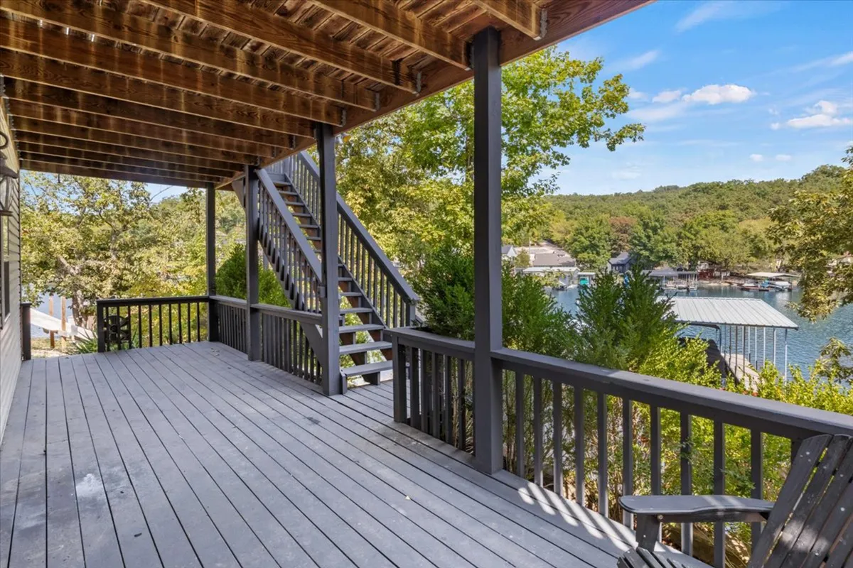 A broad view of a wooden deck overlooking Bogue Bay, bordered by trees, with the water sparkling in the background. The deck is partially shaded and open, showing stairs leading down and suggesting easy lake access. The scene captures people relaxing outdoors and hints at evening gatherings as the sun sets over the water.