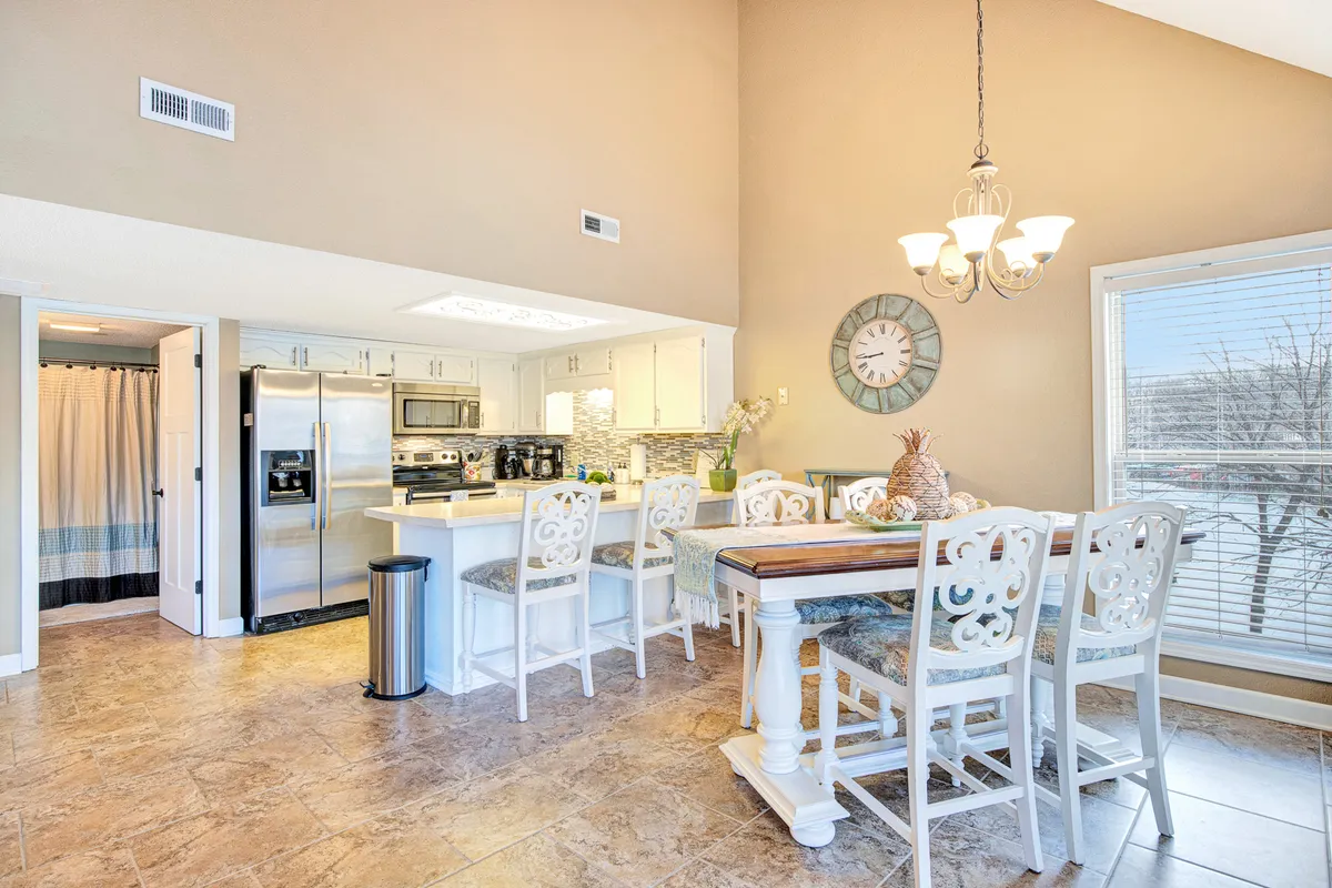 A modern, inviting dining area with a large dining table, a kitchen island in the background, a striking chandelier overhead, and a decorative wall clock. The environment suggests family meals, entertaining, and a well-cared-for home.