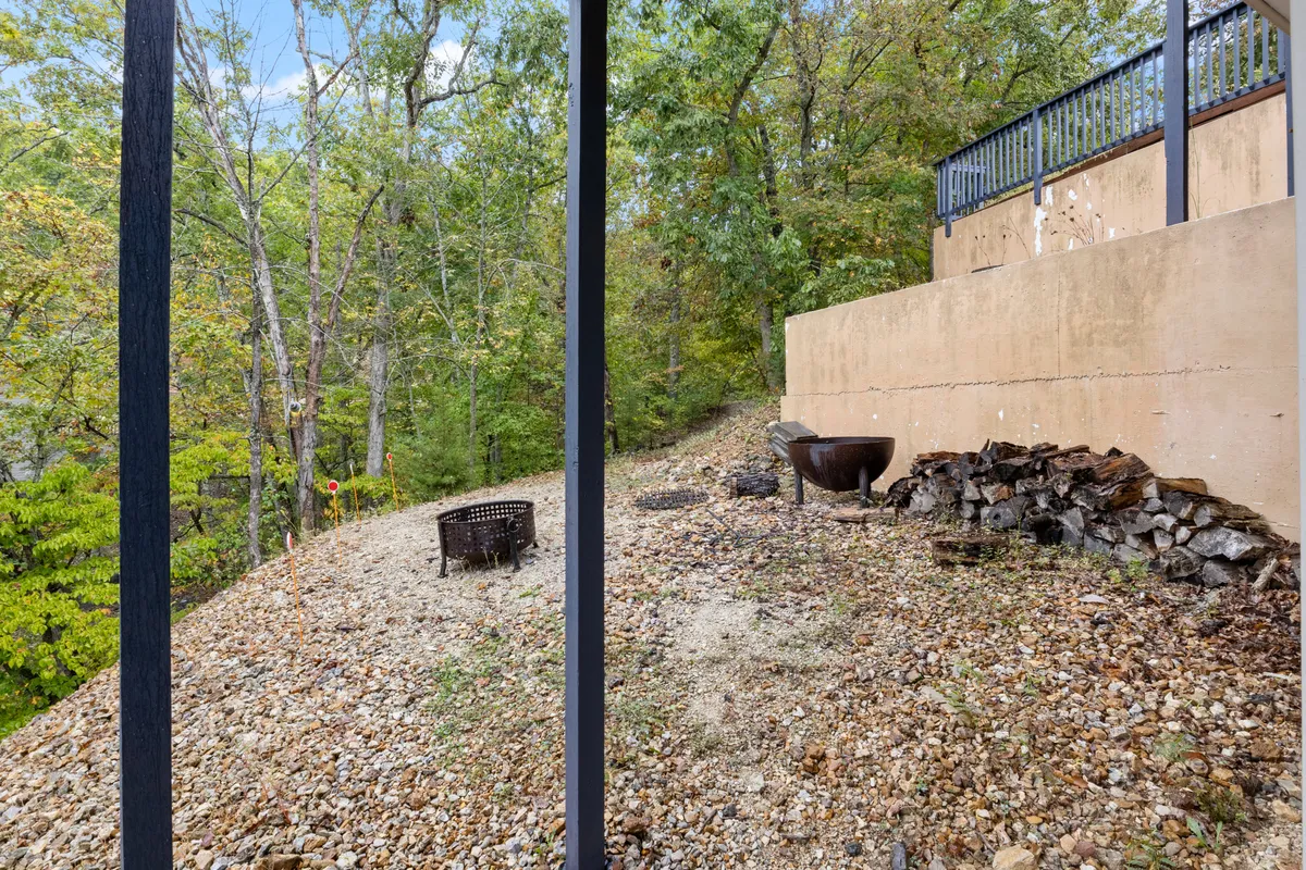 The image features a cozy fire pit area surrounded by seating and natural woodland, inviting guests to gather around for conversation, warmth, and stargazing. The wood pile and rustic setting reinforce the inviting, communal feel.